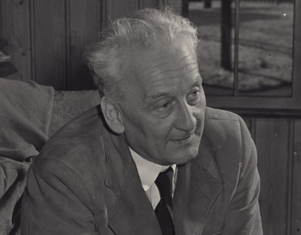 Albert sits on a chair, leaning forward with his elbows on his legs and hands clasped.