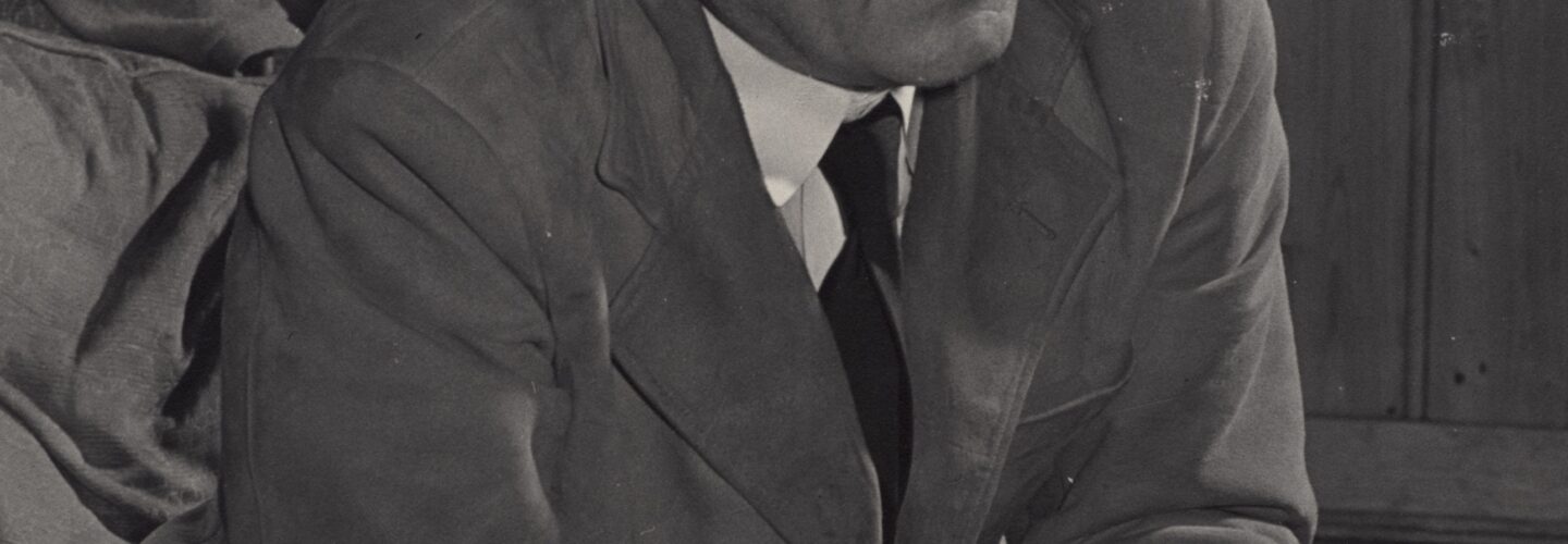 Albert sits on a chair, leaning forward with his elbows on his legs and hands clasped.