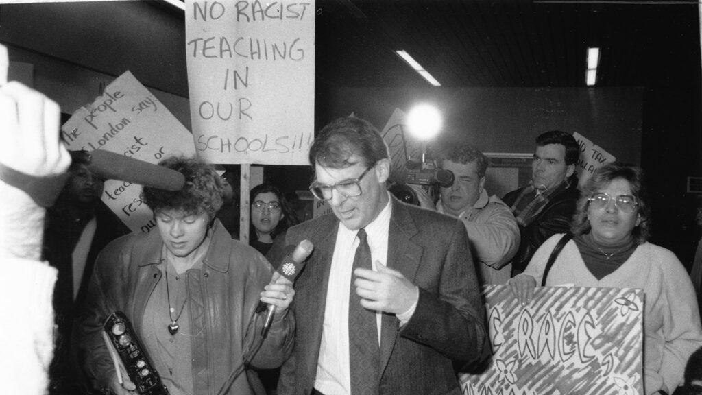 Black and white photo of man in suit surrounded by reporters and sign waving protestors