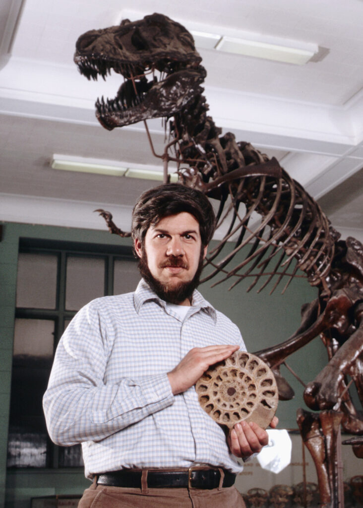 color photograph of man standing in front of dinosaur exhibit holding a fossil