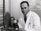 Charles Drew is seated at a counter wearing a lab coat and touching a microscope