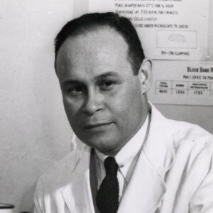 Charles Drew is seated at a counter wearing a lab coat and touching a microscope