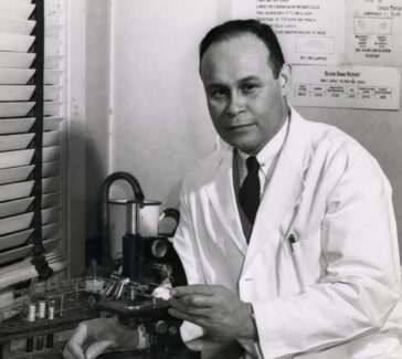 Charles Drew is seated at a counter wearing a lab coat and touching a microscope