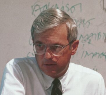Fuller in a white button up and tie, wearing glasses and sitting in front of a dry erase board