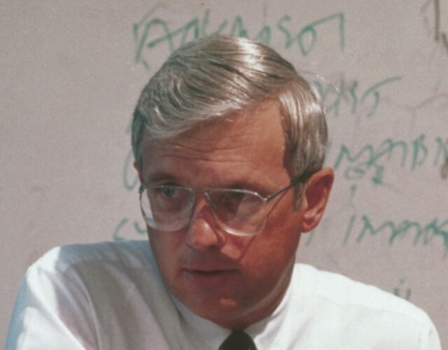 Fuller in a white button up and tie, wearing glasses and sitting in front of a dry erase board