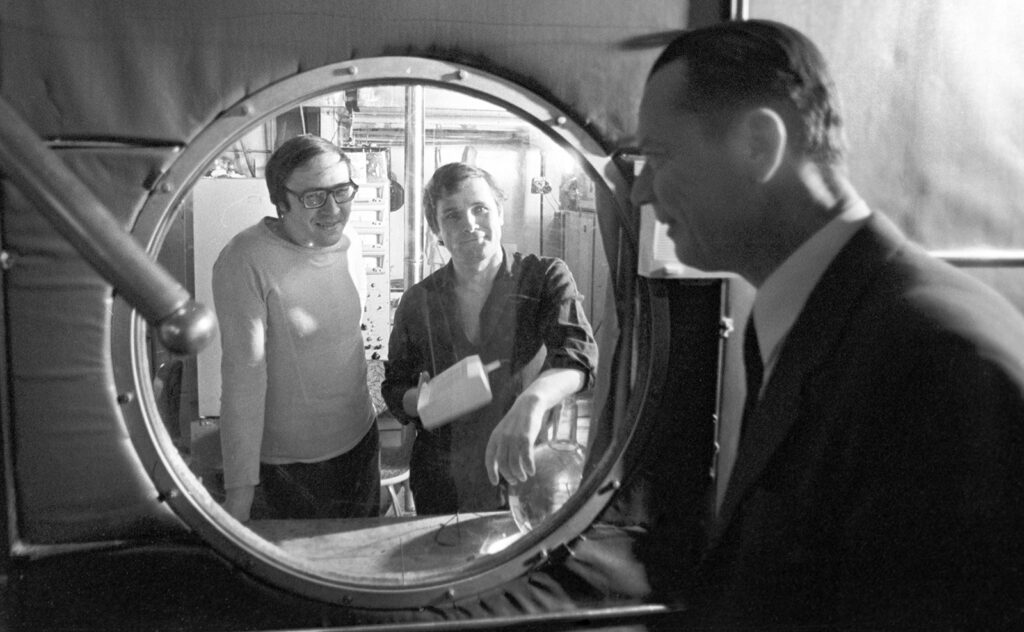 Photo of two men behind porthole talking to an older man