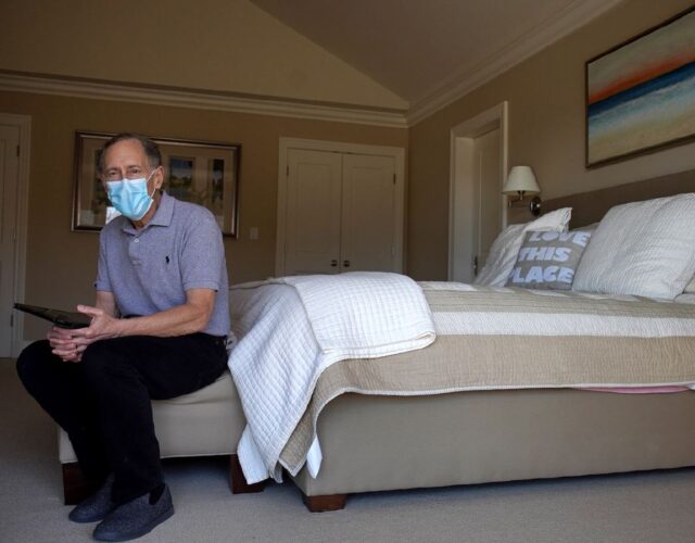 Photograph of Robert Langer sitting at the foot of a bed, wearing a surgical mask.