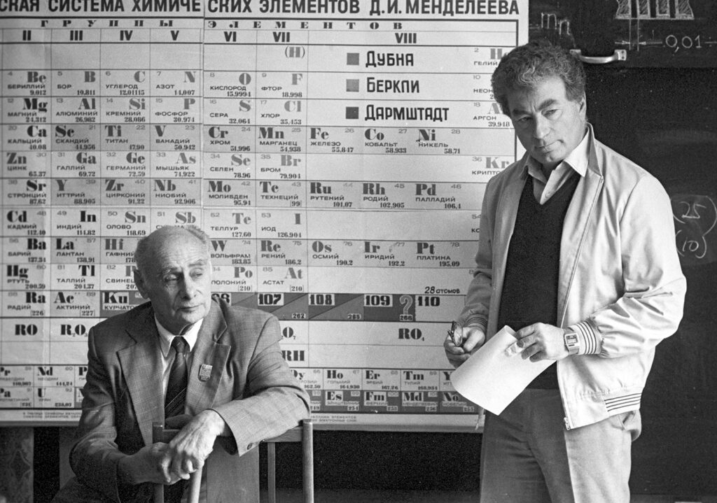 Two men (one seated) standing before a periodic table with Cyrillic characters
