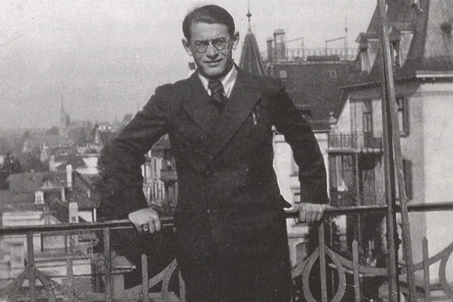 a black and white photograph of a man with  glasses in a suit and tie, leaning up against a railing with his hand propped on the railing for support. 