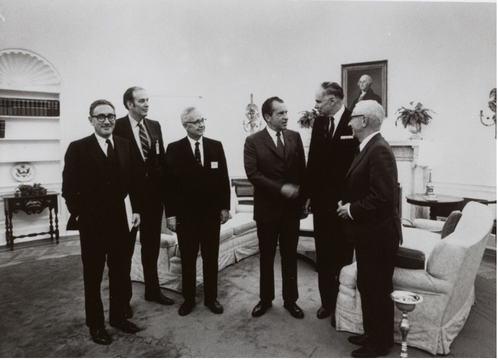 Six men in suits, including Seaborg and Nixon, standing around in a living room-styled space. Glenn Seaborg and Richard Nixon are shaking hands.