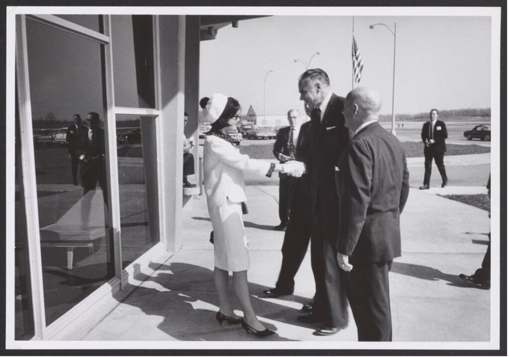 Black and white photo of Glenn Seaborg in a suit greeting First Lady Jacqueline Kennedy in a white two-piece blazer and skirt. Men in suits stand around them.