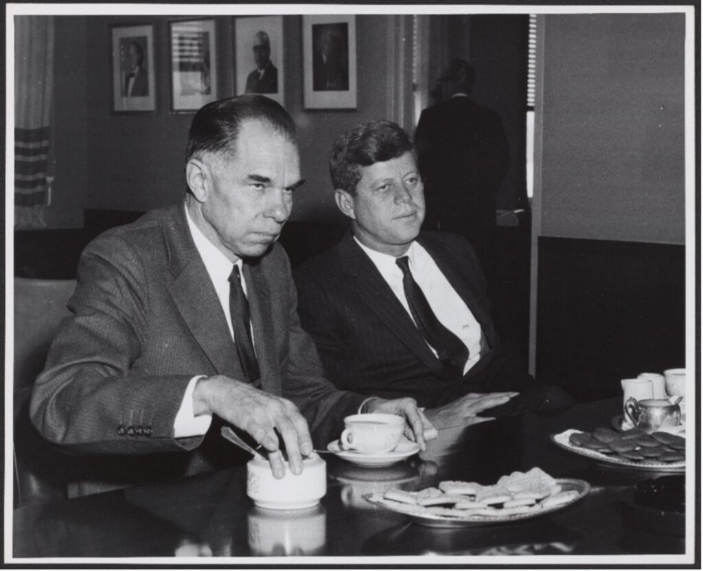 President John F. Kennedy and Glenn Seaborg are wearing suits and dark ties. They are seated at a table with coffee and cookies.