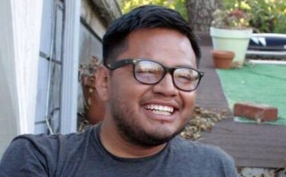 Rigo sitting on steps and leaning back on his left arm. He is wearing black glasses and a grey T-shirt. He is smiling.