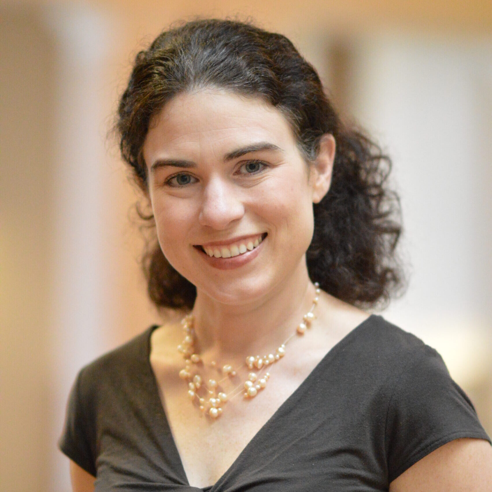 Clare smiling with her curly hair pulled back wearing a brown shirt and pearl necklace