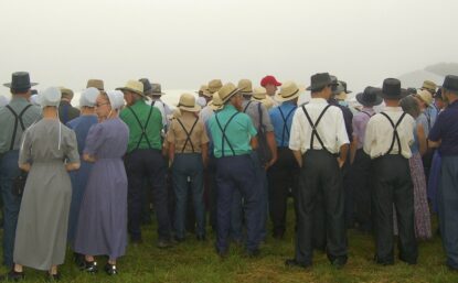 A photo of a group of Amish men and women from the back. They are standing in a field.