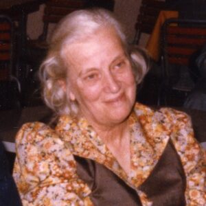 Dorothy Hodgkin in 1991 is shown seated and smiling. Her hair is half up and she is wearing a floral dress with brown panels.