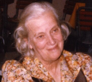 Dorothy Hodgkin in 1991 is shown seated and smiling. Her hair is half up and she is wearing a floral dress with brown panels.