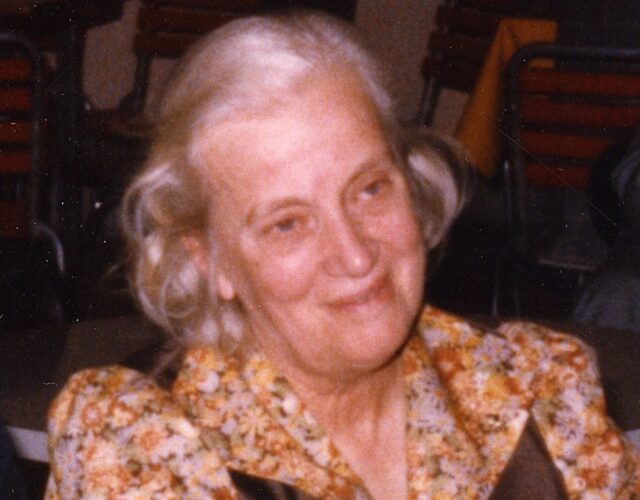 Dorothy Hodgkin in 1991 is shown seated and smiling. Her hair is half up and she is wearing a floral dress with brown panels.