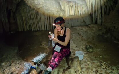 Photograph of scientist Gabriela Serrato Marks wearing a headlamp in a cave, surrounded by samples in plastic bags.