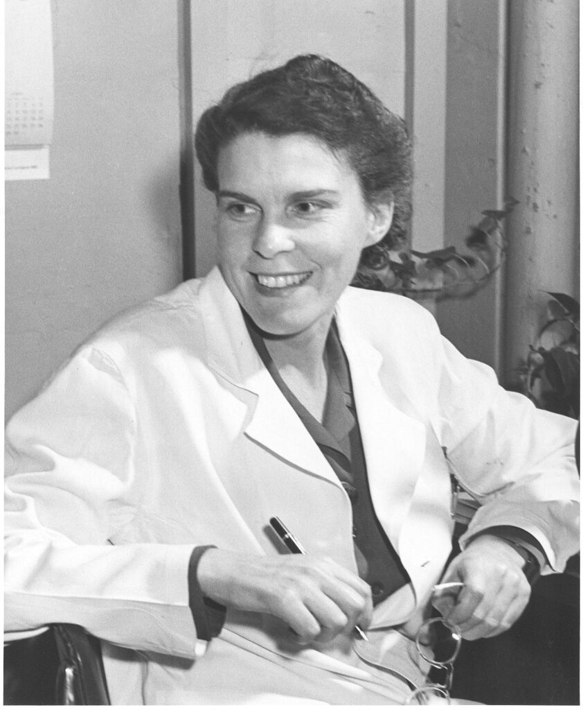 A woman in a white lab coat sits in a chair smiling