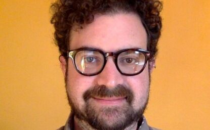 Jonathan with curly hair and glasses wearing a grey shirt. There is an orange background.