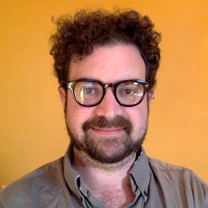 Jonathan with curly hair and glasses wearing a grey shirt. There is an orange background.