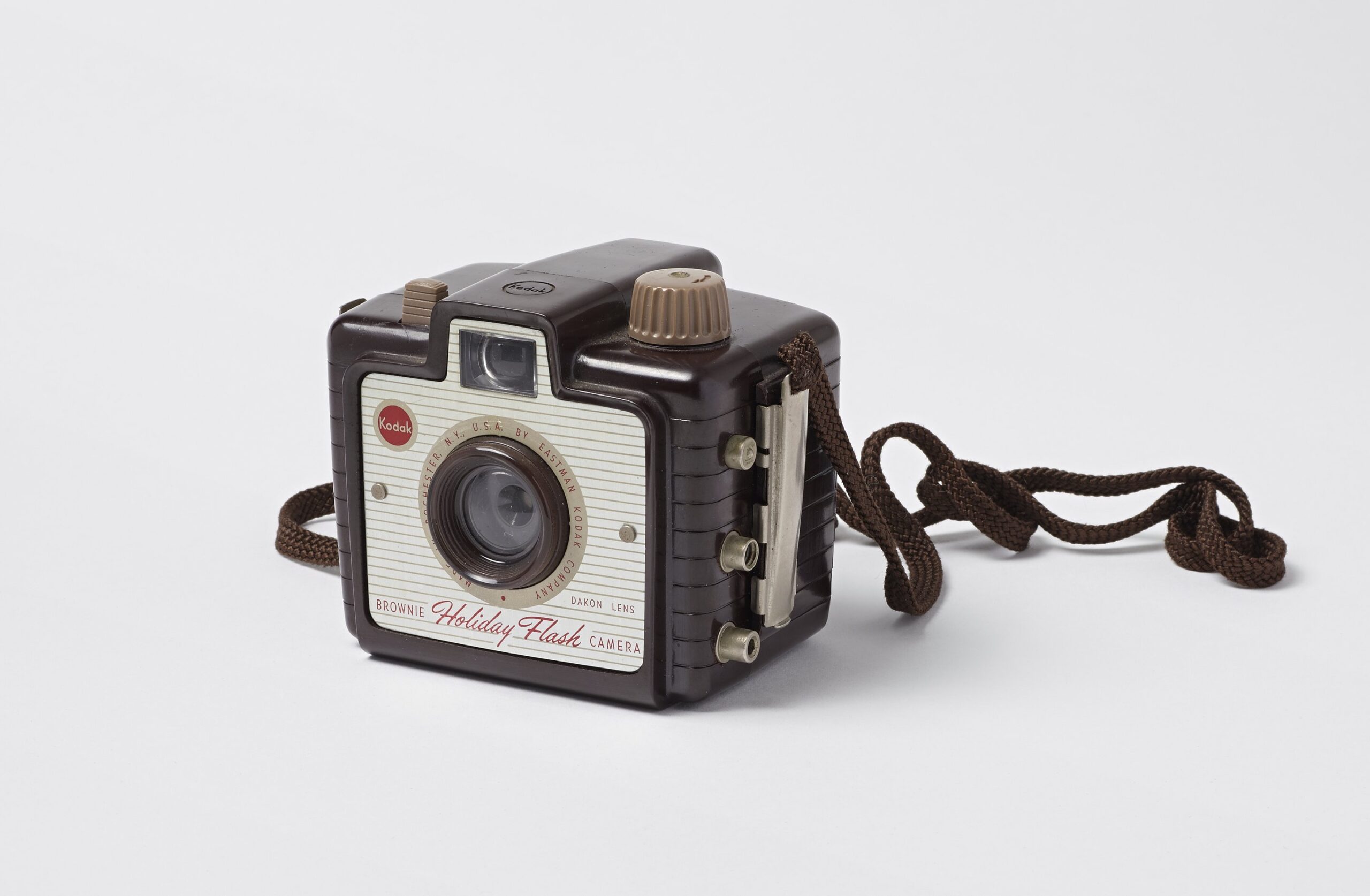 Kodak brand Brownie Holiday Flash camera; Dakon plastic lens with rotary shutter; molded brown and tan Bakelite body; three metal sockets on right side of the camera body meant for mounting flash bulb; braided cord carrying strap.