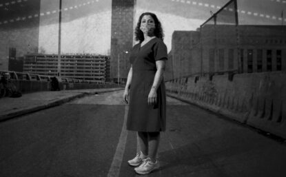 Black and white photograph of nurse Linda Ruggiero wearing a face mask, standing outside on a bridge.