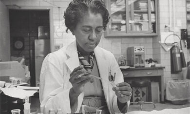 An image of Marie Maynard Daly, a black woman scientist in a lab coat works with test tubes in a laboratory.