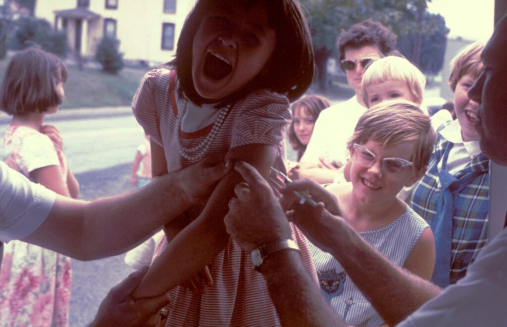 A young girl in a pink, striped dress is injected with a vaccine. She is yelling and laughing while children behind her watch.