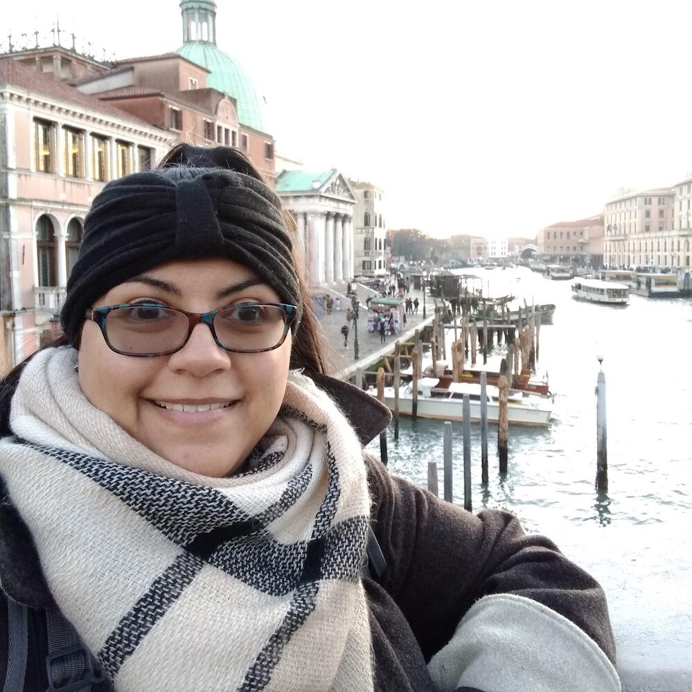 Nyomi smiles and wears a thick black headband and plaid scarf. There is a canal in the background.