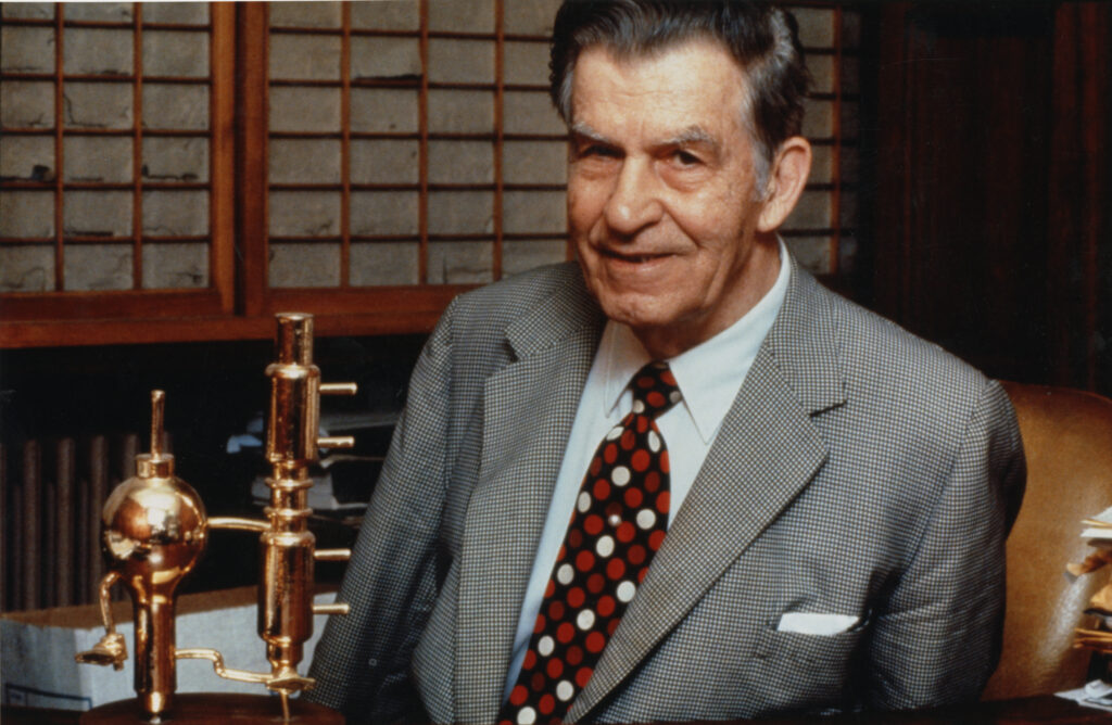 Donald Othmer wearing a suit and tie, with a gold-plated Othmer still in the background. 