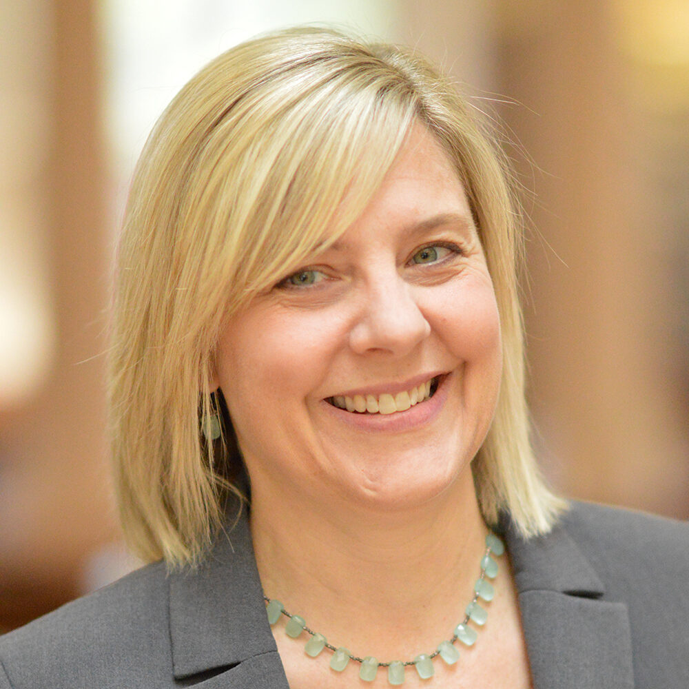 Resa in a grey blazer wearing a green necklace, smiling