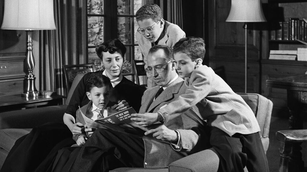 A man sitting on a loveseat with his wife, reading Time Magazine; around them are their young sons.