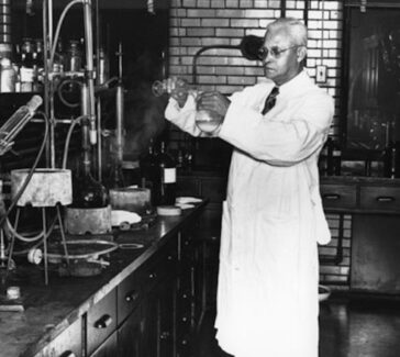 St. Elmo Brady in a chemistry lab at Fisk University. He is pouring a liquid from one beaker into another. There are counters of lab equipment surrounding him.