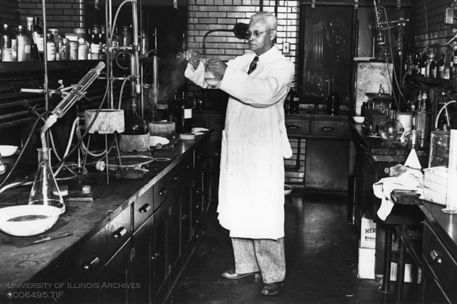 St. Elmo Brady in a chemistry lab at Fisk University. He is pouring a liquid from one beaker into another. There are counters of lab equipment surrounding him.
