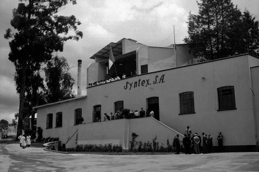 Black and white photo of a building with "Syntex S.A." on the facade. There are people standing outside of the building.