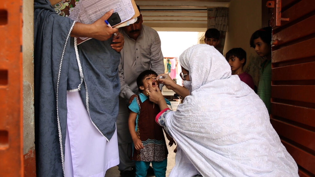 A woman holds a small boy’s mouth open while administering a vaccine. Two men stand behind him, holding him still.