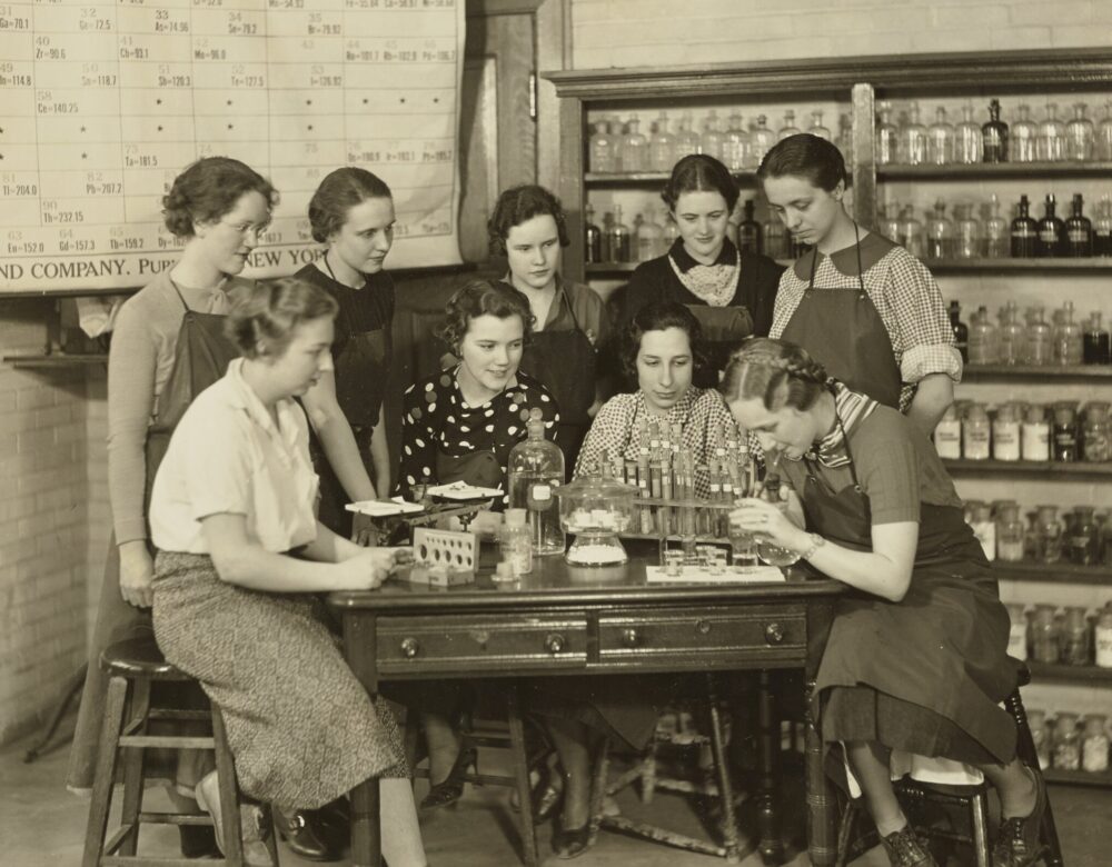 group of women standing around a lab table with periodic table in background