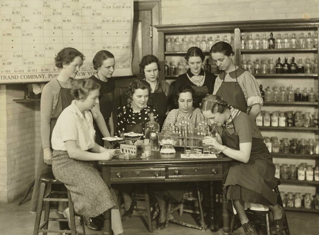 group of women standing around a lab table with periodic table in background