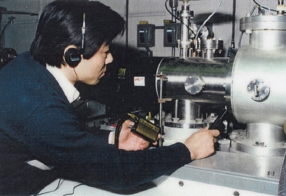 A man in a blue sweater wearing headphones holds a microphone towards a large metal piece of scientific equipment.