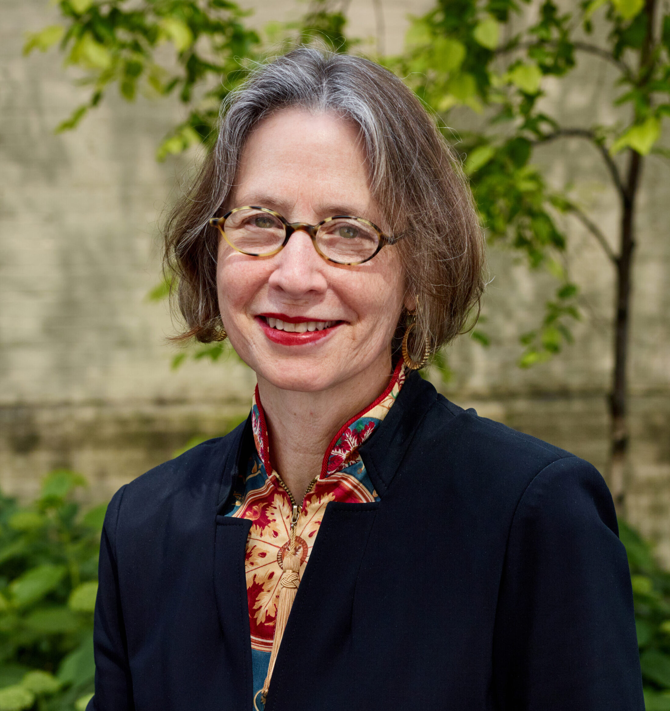 Grace outdoors, smiling, wearing glasses and a colorful blouse under a navy blue blazer