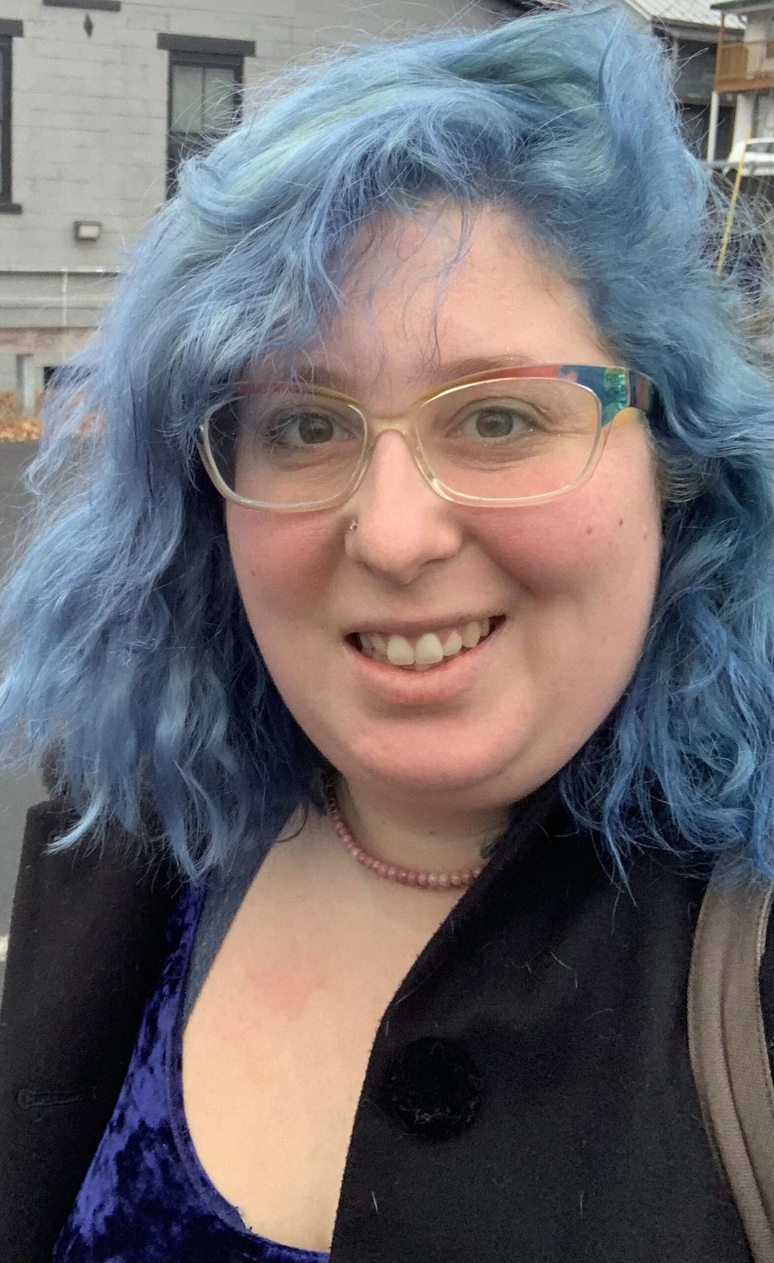 Jennifer Lupu, outdoors in an urban location, glasses with multicolor frame, smiling, hair dyed blue, purple velour top under black jacket, coral necklace