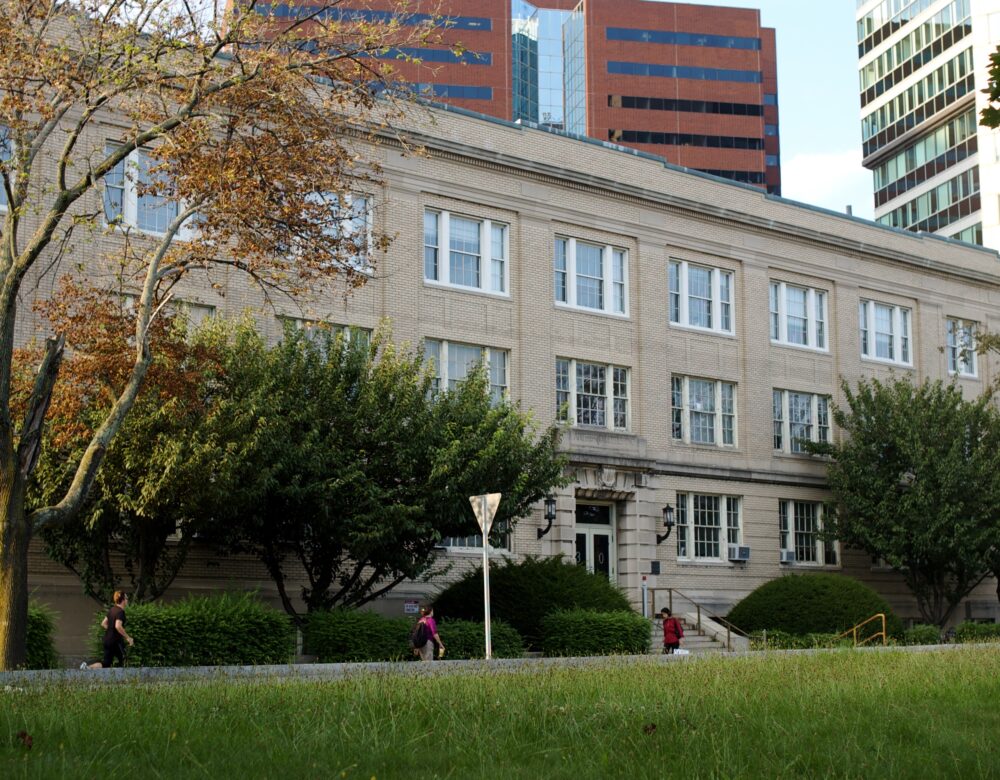 Sand-colored brick building, three stories, grass and people walking in front, high-rise office buildings behind