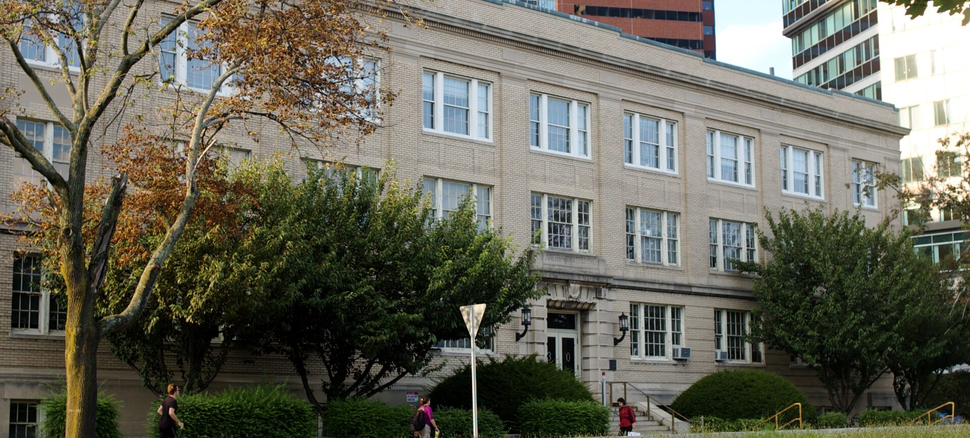 Sand-colored brick building, three stories, grass and people walking in front, high-rise office buildings behind