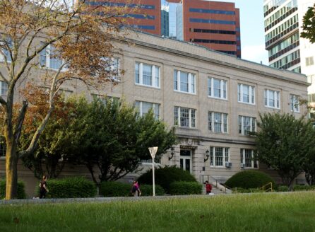 Sand-colored brick building, three stories, grass and people walking in front, high-rise office buildings behind