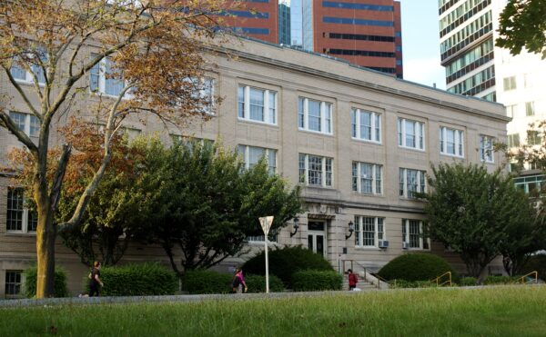 Sand-colored brick building, three stories, grass and people walking in front, high-rise office buildings behind