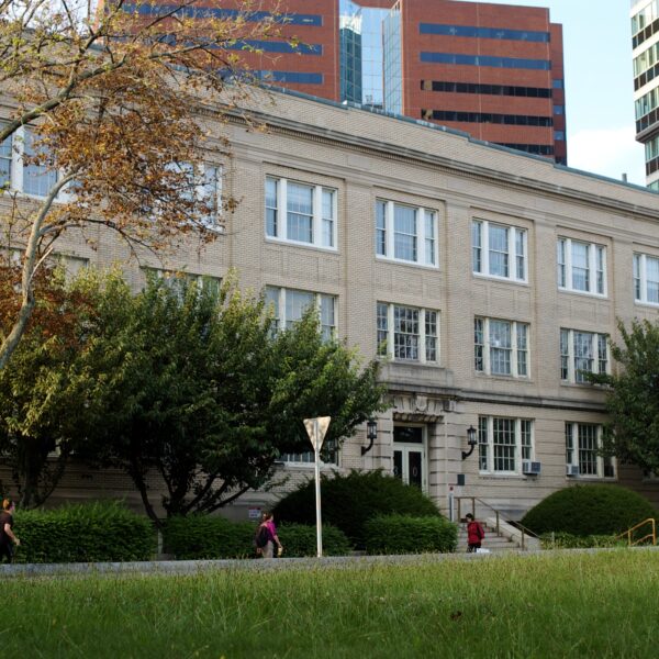 Sand-colored brick building, three stories, grass and people walking in front, high-rise office buildings behind