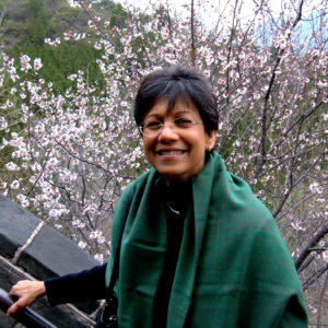 Uma Chowdhry wearing green shawl and glasses, hand on a rail, cherry blossoms in background