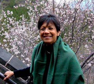 Uma Chowdhry wearing green shawl and glasses, hand on a rail, cherry blossoms in background